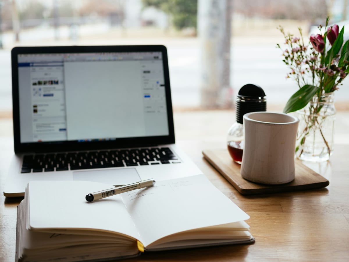 photo of a laptop and a notebook with a pen and a mug of coffee on a desk